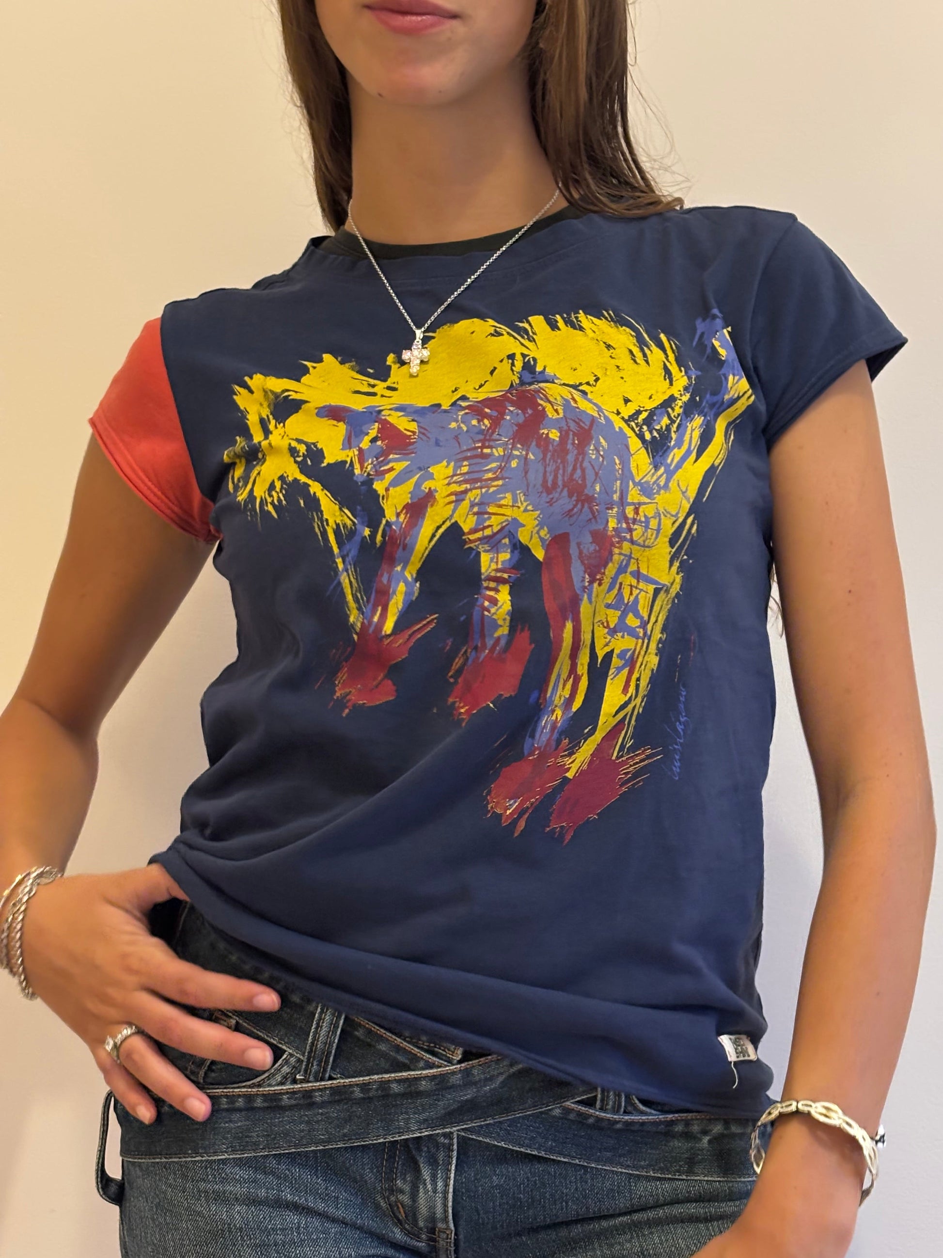 A woman wearing our Cats tee, a navy blue graphic t-shirt with a colorful abstract cat print and dark wash jeans, shown  against a plain beige wall.