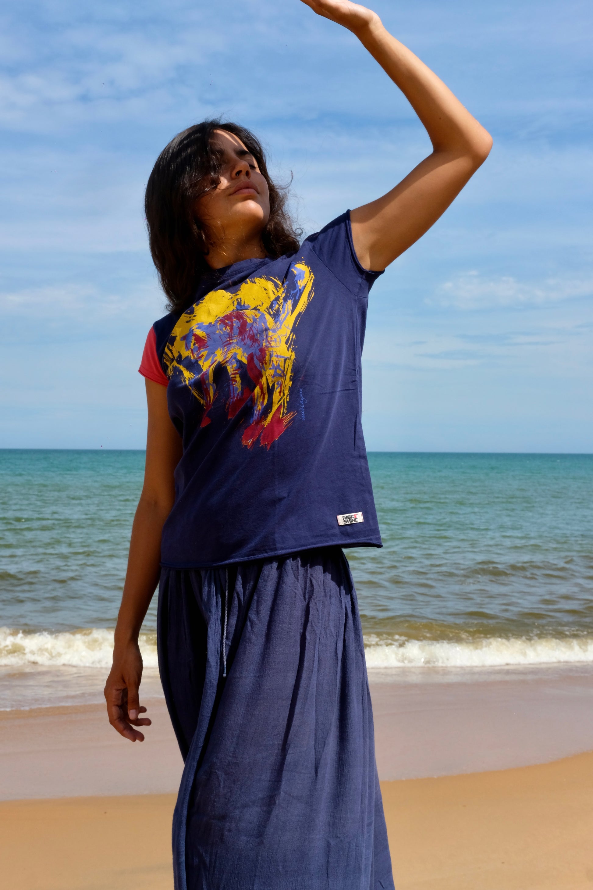 A woman wearing our Cats tee, a navy blue graphic t-shirt with a colorful abstract cat print and matching navy blue skirt stands on a sandy beach with the ocean and blue sky in the background.