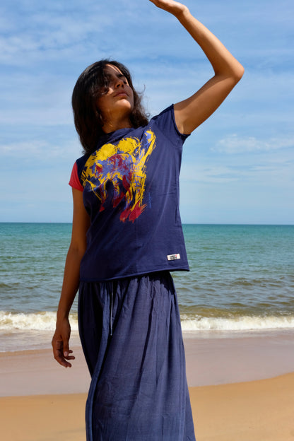 A woman wearing our Cats tee, a navy blue graphic t-shirt with a colorful abstract cat print and matching navy blue skirt stands on a sandy beach with the ocean and blue sky in the background.