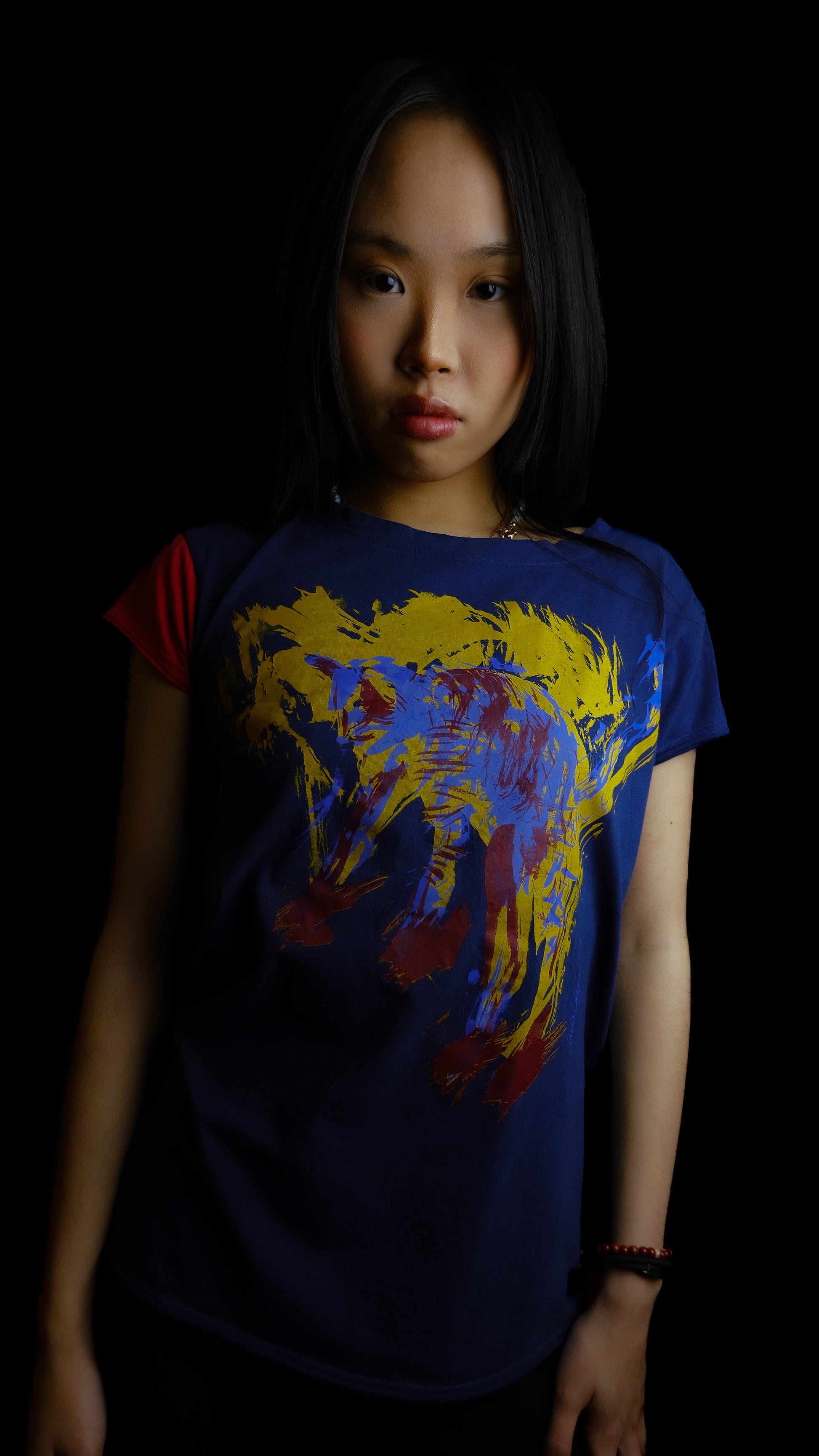 A young woman with long black hair wearing our Cats tee, a dark navy blue graphic t-shirt with a colorful abstract cat print, posed against a solid black background.