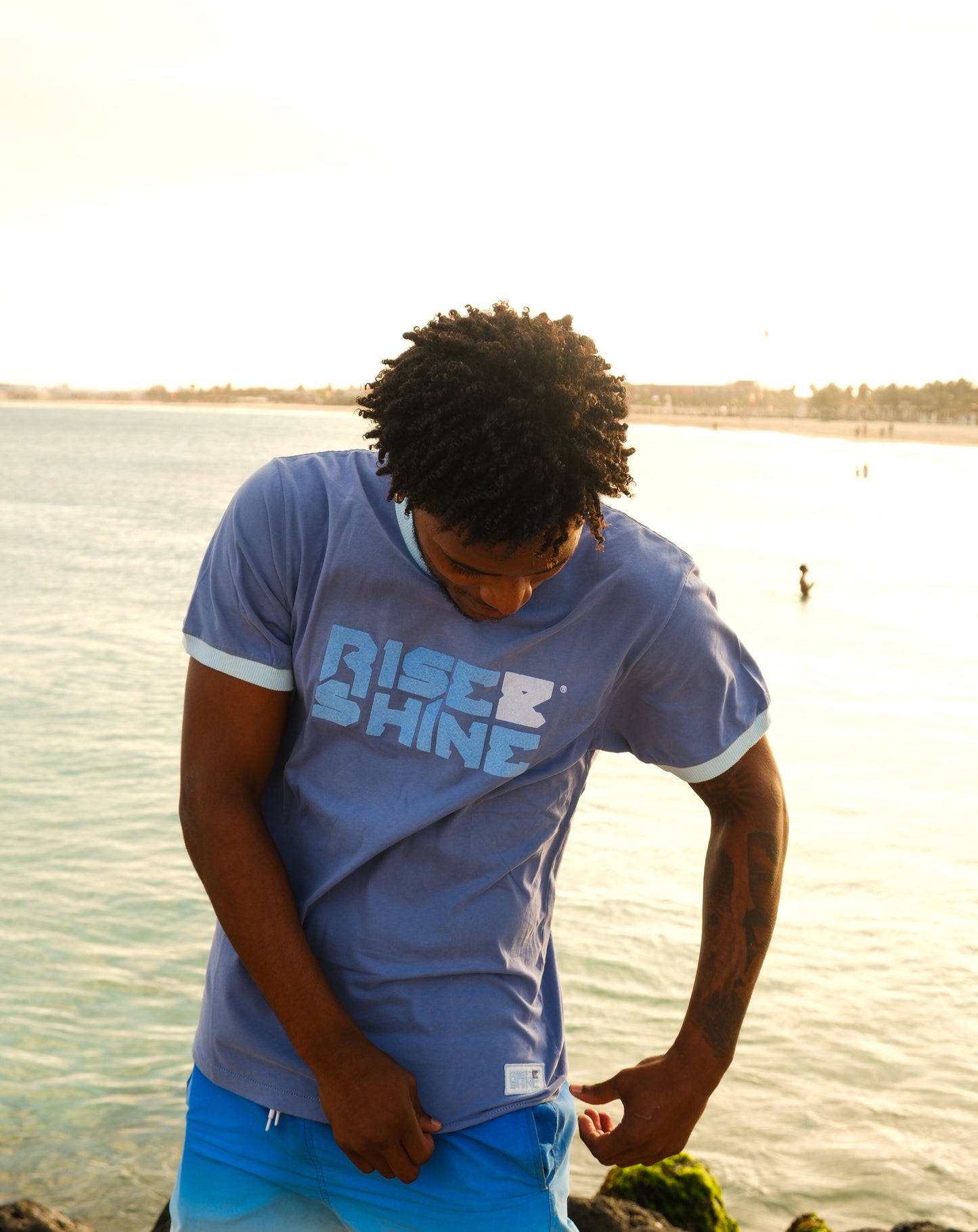 Man wearing the signature blue Rise & Shine t-shirt on a pier by the beach in Sal, Cabo Verde.