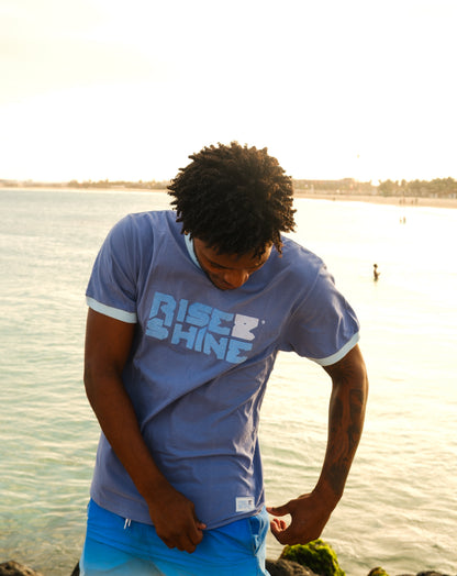 Man wearing the signature blue Rise & Shine t-shirt on a pier by the beach in Sal, Cabo Verde.