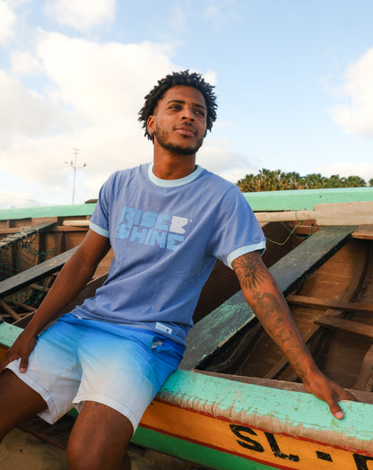Man wearing the signature blue T-shirt with the Rise & Shine logo sitting on a wooden boat on the beach in Sal, Cabo Verde.