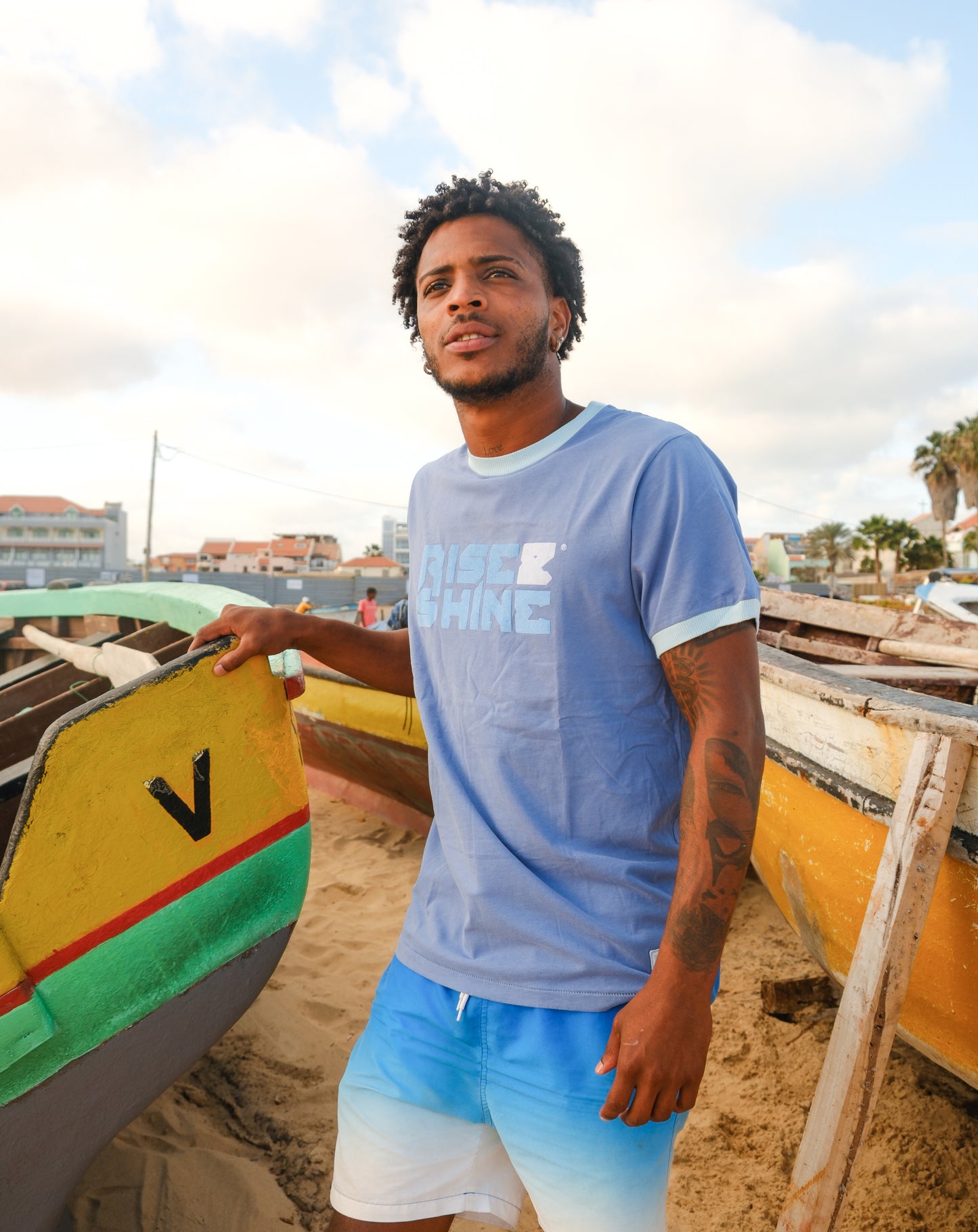 Man wearing the signature blue T-shirt with the Rise & Shine logo standing next to a wooden boat on the beach in Sal, Cabo Verde.